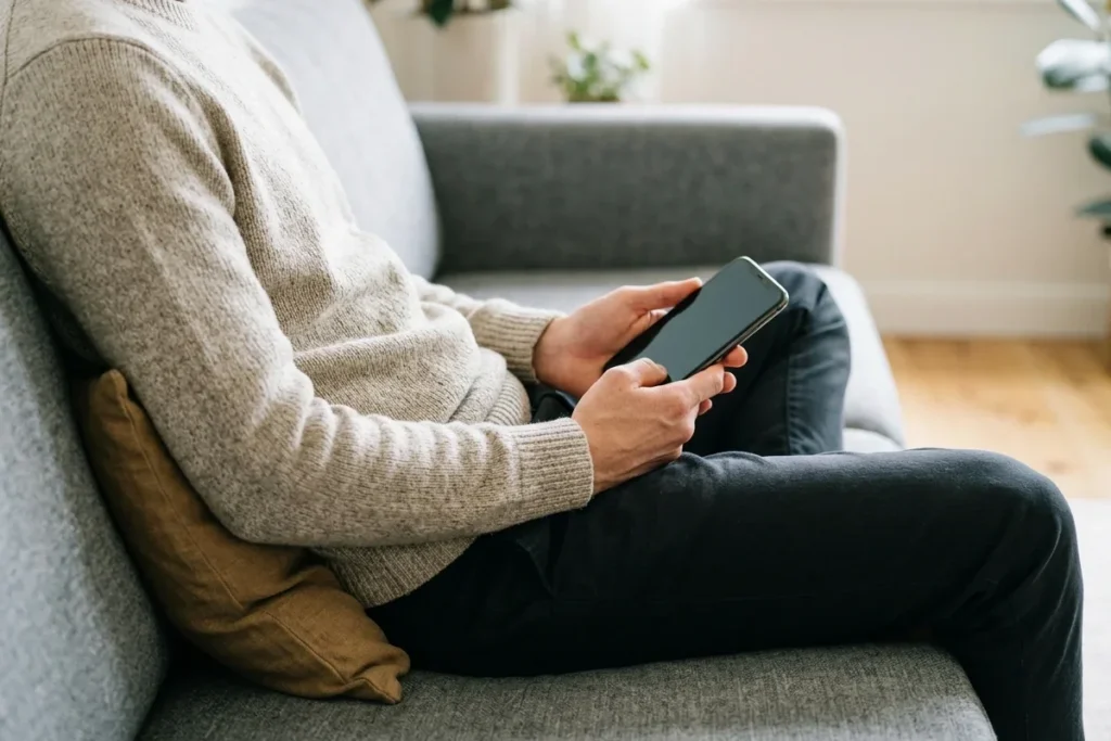 Person sitting on a sofa with a cushion behind the lower back, holding a phone