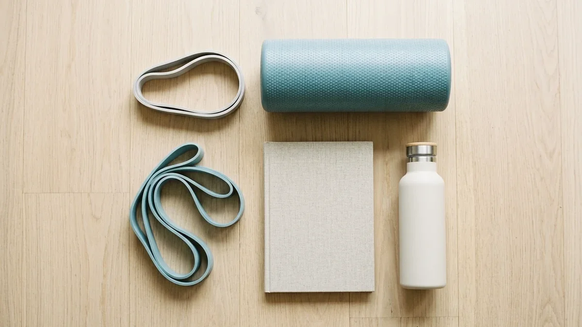 Resistance bands, a foam roller, a notebook and a water bottle laid out on a wooden floor